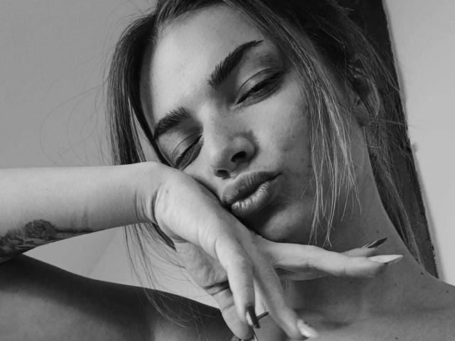 Ingrids Gesicht mit halb geschlossenen Augen, leicht geschürzten Lippen und einer Hand, die ihr Kinn berührt, wobei ein Tattoo an ihrem Handgelenk sichtbar ist. Schwarz-weiß-Foto, mit sichtlichem Eyeliner und langen Nägeln..