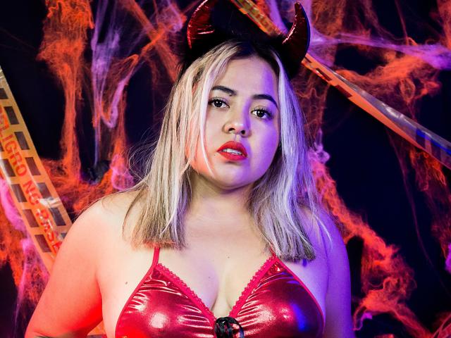 GraceJansen with blonde hair, red devil horns, and shiny red bra, showing her tits, red lipstick, and serious expression. Background has red smoke and film strip.
