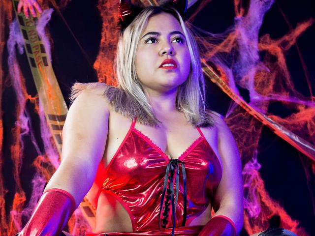 GraceJansen with blonde hair, red devil horns, and a shiny red latex bra, showing her tits, wearing red gloves, looking up with a slightly open mouth in a dark setting.
