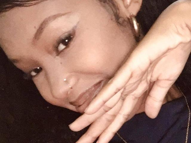 Stephah's face with dark brown skin, small nose piercing, and gold hoop earrings, smiling slightly, holding her chin with her hand. Her eyebrows are thick and arched.