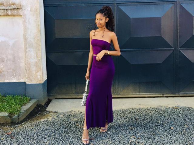 Stephah is standing in a purple strapless dress, holding a silver purse, with dark curly hair in a high ponytail, wearing clear heels, on a gravel path.
