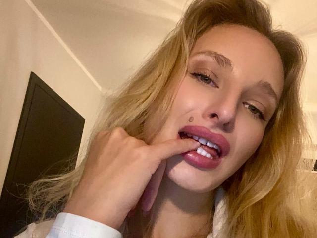 Marilyn, avec ses cheveux blonds, légèrement ondulés, mordant sa lèvre supérieure rose, montrant ses dents blanches, son doigt sur son menton, portant une chemise blanche, sur une photo de soi..