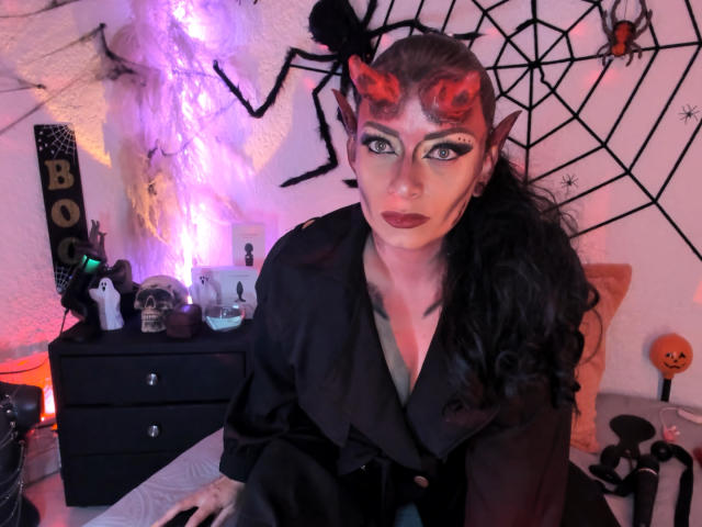 JeaninexScott with dark makeup, red devil horns, and black eyeliner, wearing a black satin robe, in a spooky room with purple and orange lights, and Halloween decorations.
