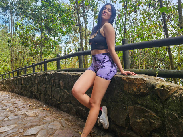 Nico Coleman, vestindo shorts roxos e um top esportivo preto, apoiada em uma parede de pedra, exibindo suas nádegas, usando tênis brancos, em pé em um caminho de pedras em uma floresta..