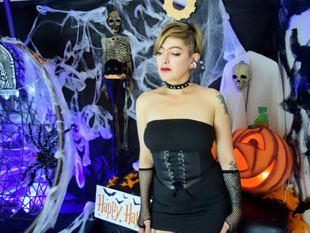 MaferCougar with short blonde hair, black strapless dress, corset, fishnet arms, choker, tattoos, standing in Halloween setup, skeletons, pumpkin, and spooky decorations. Boobs, dress, corset, choker.