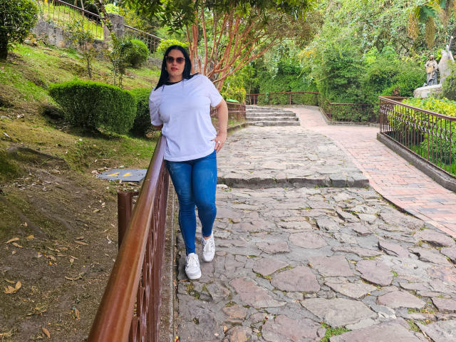 Liza Russell está de pie, apoyada en una barandilla de madera, vestida con una camisa blanca, jeans azules y zapatillas blancas, con cabello oscuro y gafas de sol, en un camino de piedra..