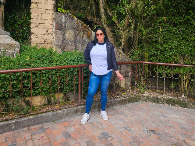 LizaRussell has medium tits, wears a white tee, blue jeans, and a black jacket with white sneakers, standing with a hand on her hip, looking happy.