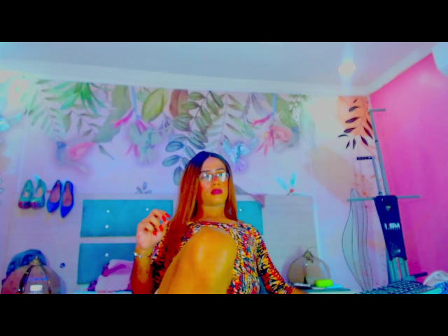 MiaFoxTs with long red hair, wearing glasses and a colorful patterned top, holding a knee in a brightly lit room, neon pink and blue lighting.