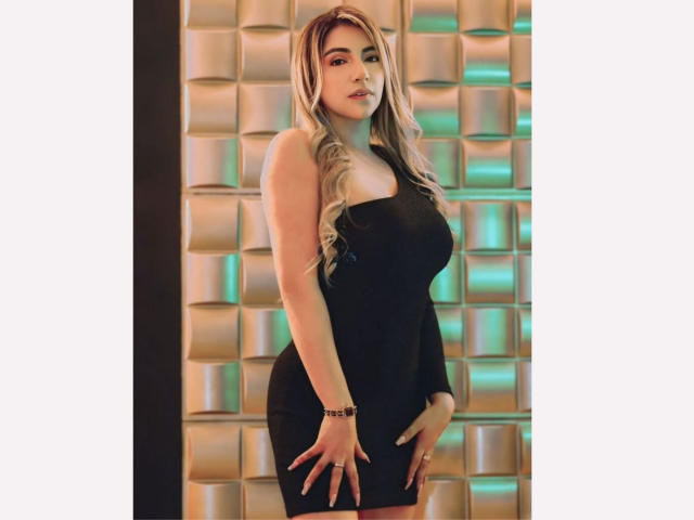 CarolinaaGrey, with big tits and a curvy ass, wears a tight black dress, long blonde hair, and a bracelet, standing with hands on her thighs.