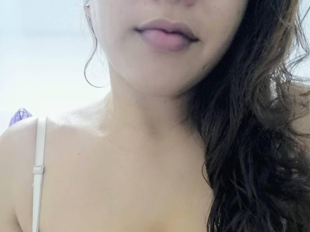 AshleyCurvyHot's face, showing slightly parted pink lips, wavy brown hair, and a white strap from a dress on her shoulder. Her skin is light and smooth.