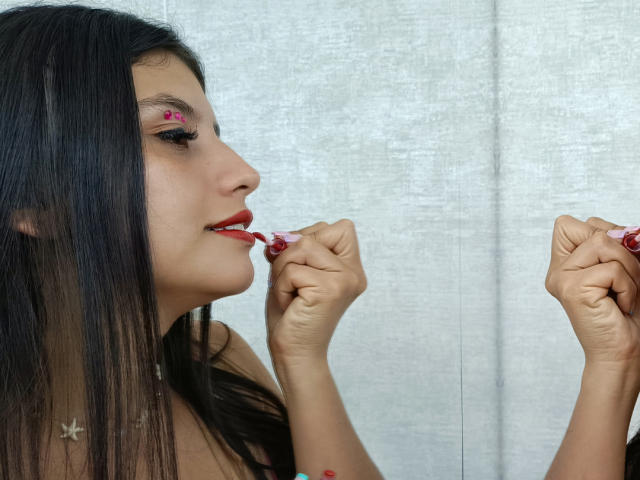 MadissonFontaine with long black hair, red lipstick, and pink nail polish, biting a pink candy in front of a mirror, showing a small red mark on her cheek.