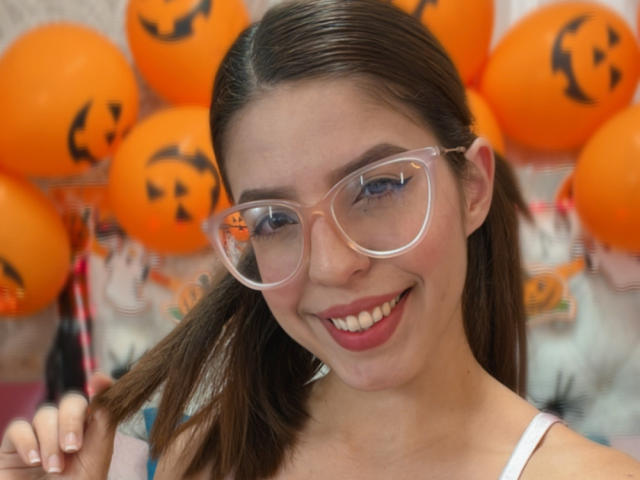 NatyBakers con gafas, sonriendo, cabello castaño, labios rosados, blusa blanca, sujetando el cabello, globos de Halloween naranjas con caras negras en el fondo..