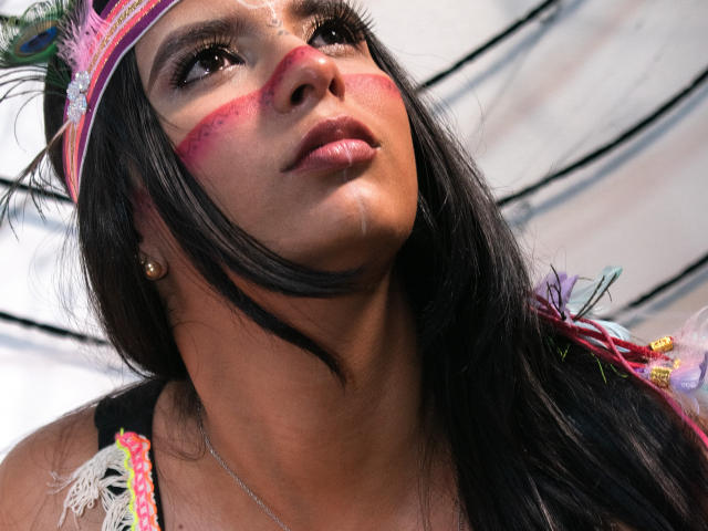 MariaSmith's face with dark hair, pink face paint, and a colorful hat, looking up with slightly parted lips. She's wearing a black top with lace and small earrings.