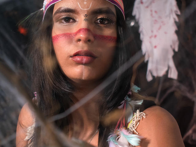 Maria Smith, com cabelo escuro, maquiagem vermelha no rosto e um ponto branco na testa, usando uma tiara rosa e uma blusa colorida com penas, com uma expressão séria através de uma janela de vidro. Seus seios estão ligeiramente visíveis..