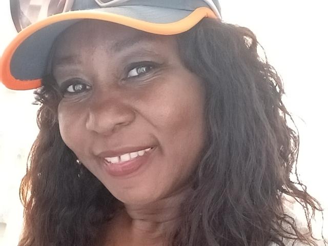 LucyLina69 with dark brown skin, wavy black hair, wearing a blue and orange baseball cap, smiling with white teeth, looking directly at the camera.