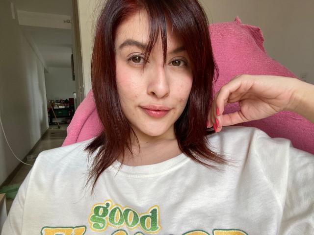 IsabellaPalacio with red hair, white shirt, and pink chair, showing a slight smile, right hand near head, light skin, subtle freckles, and brown eyes, in a relaxed selfie.