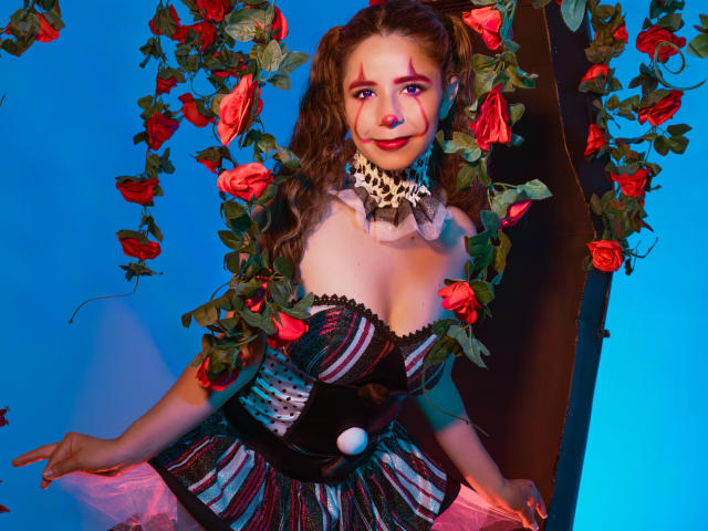 Sophie Teller, avec des roses rouges dans les cheveux, portant un corset rayé, un collier en dentelle et du tulle rose, a de jolies seins et un joli visage, sur un fond bleu..