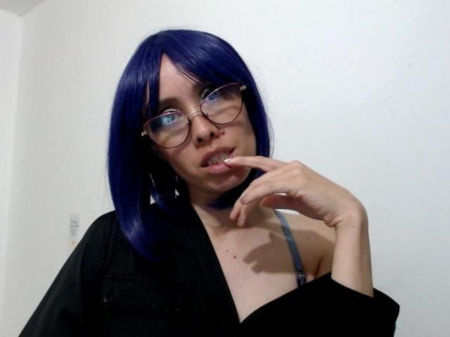 AnnaBecovka with blue bob, glasses, black shirt, and gray bra, finger near mouth, looking slightly up. Visible cleavage, pale skin, small mole on chest.
