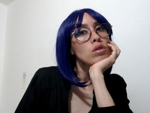 AnnaBecovka with blue bob, wearing glasses, black top, hand on cheek, looking slightly to the side, pale skin, small mole above lip, subtle makeup, white wall background.