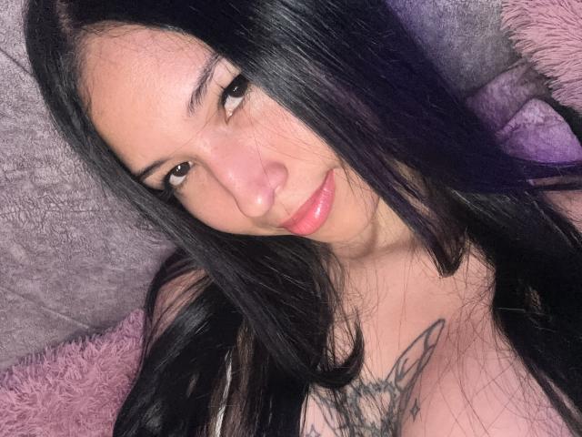 AmyAdamss with long black hair, pink lips, and a tattoo on her chest, looking at the camera with a slight smile. She has medium-sized boobs and a cute face.