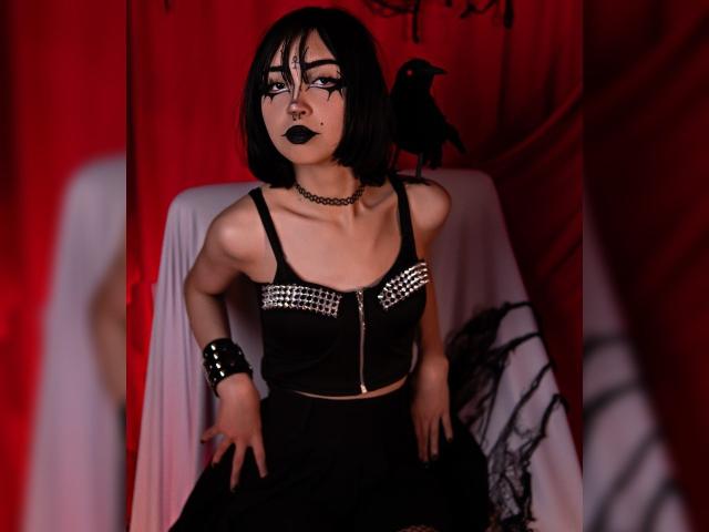 MillaHottLove69 with black bob, goth makeup, black lipstick, choker, studded top, black skirt, wrist bracelet, and black crow on chair, in red and white setting.