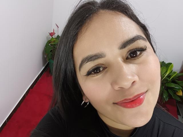 Sofiacopers has shiny brown eyes, black eyebrows, and glossy red lips. Her black hair is straight, and she's wearing a black shirt with small earrings.