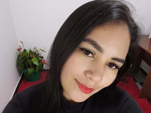 Sofiacopers has dark straight hair, brown eyes with long eyelashes, red lipstick, and a black shirt, smiling slightly. Her face is centered, with a green plant in the background.