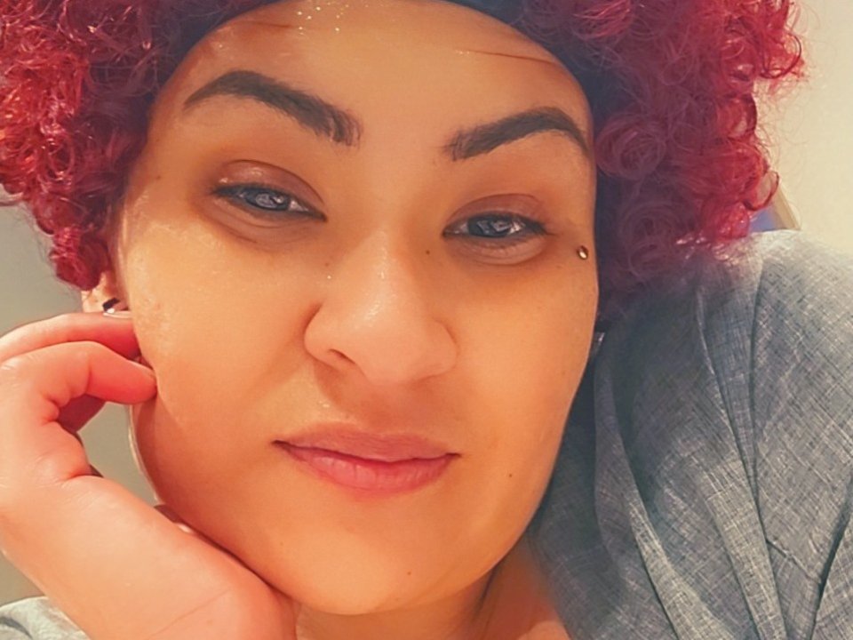 SweetPeach69s with curly red hair, thick eyebrows, small beauty mark on right cheek, light brown skin, wearing grey shirt, looking at camera with slight smile, hand near face.