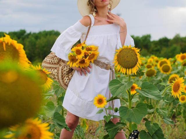 AdrianaSea stands in a sunflower field, wearing a white dress, sunhat, and holding a sunflower bouquet, with a woven bag, and her tits slightly visible.