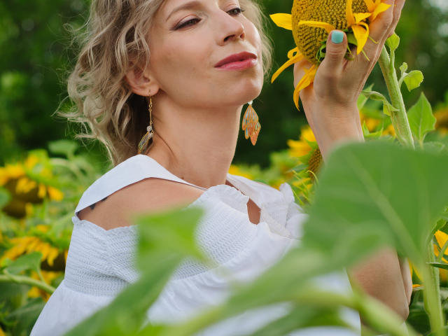 AdrianaSea with wavy blonde hair, white dress, and flower earrings, holding a sunflower, smiling, showing her pink-lipped, slightly upturned nose, and blue-painted nails.