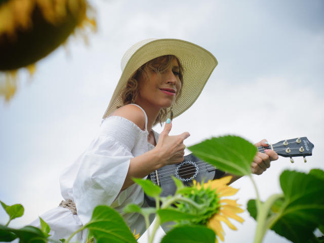 AdrianaSea with curly blonde hair, wearing a white off-shoulder top and sun hat, playing a small black guitar, surrounded by sunflowers, smiling gently.