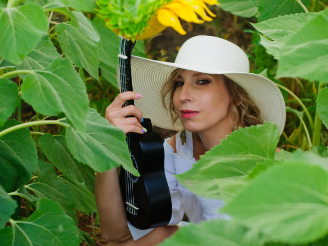 AdrianaSea with curly blonde hair, pink lipstick, white hat, holding black ukulele, surrounded by sunflowers and green leaves, looking seductive with a slight smirk.