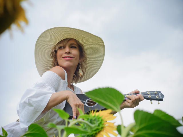 AdrianaSea with wavy brown hair, white off-shoulder top, and large sunhat, holding a black ukulele, smiling, surrounded by sunflowers and green leaves, blue sky above.
