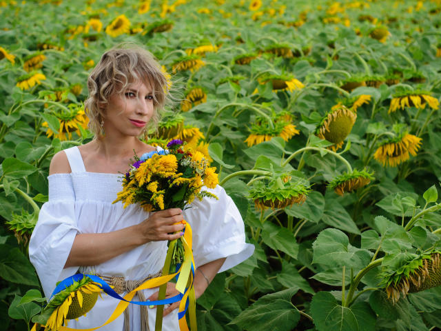 AdrianaSea with curly blonde hair, wearing a white off-shoulder dress, holding a bouquet of yellow and purple flowers, standing in a sunflower field, her tits slightly visible.