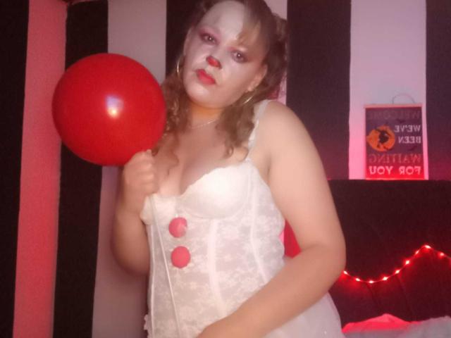 AmandaHotL with painted white face, red nose, and lipstick, holding red balloon, wearing white lace corset with pink buttons, in dimly lit room, showing her big boobs.