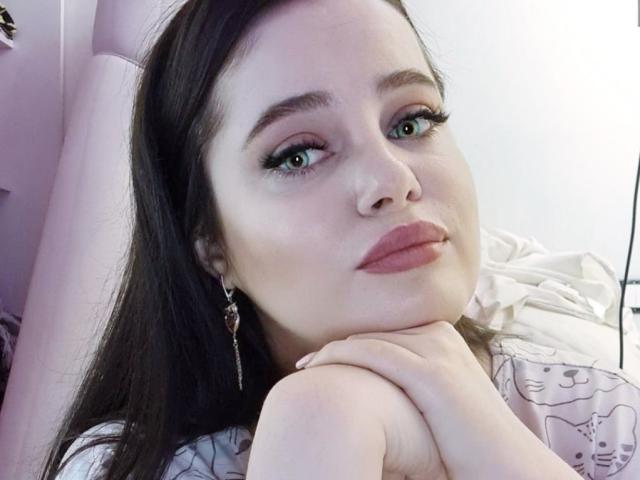 Marizel's face, looking up with green eyes, dark brown hair, pink lipstick, and a slight smile, resting her chin on her knee, wearing dangling earrings, in a pink chair.