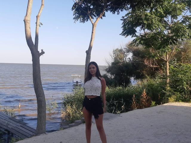 TanyaDevon stands on a sandy path with two trees, wearing a white lace top and black high-waisted shorts, showing her ass and long brown hair in a sunny beach setting.