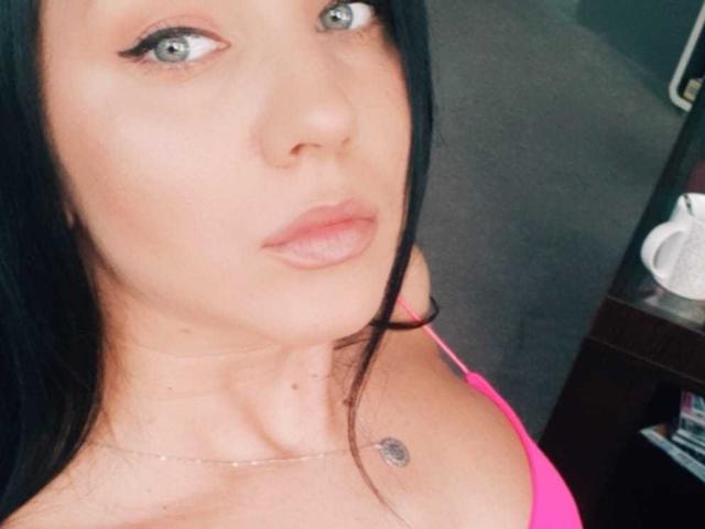 TanyaDevon has pale skin, blue eyes, and pink lipstick. She's wearing a pink top, showing her small tits and a tiny tattoo near her collarbone. Her black hair is down.
