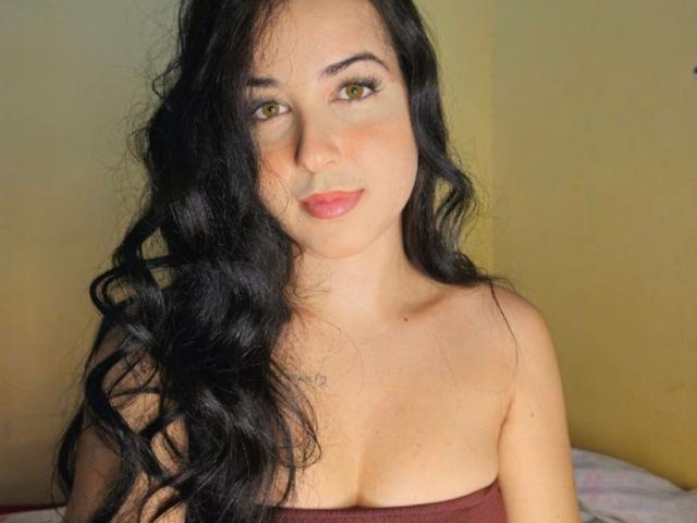 EsmeraldaXx has long, wavy black hair, yellow eyes, and is wearing a strapless brown top, showing her tits and a small tattoo on her chest. She's smiling softly.