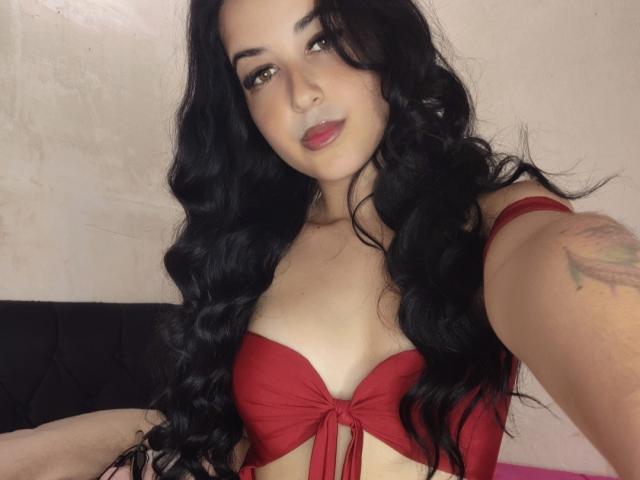 EsmeraldaXx with long black wavy hair, red bra, and tattoo on her right arm, showing off her tits, slightly parted red lips, and a sexy selfie look.