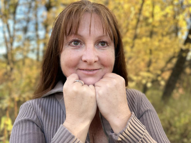 Vanessa, con capelli castani, occhi verdi, mani vicine al mento, indossa una giacca grigia a coste con zip e un anello alla mano destra, sorride dolcemente in una foresta autunnale..