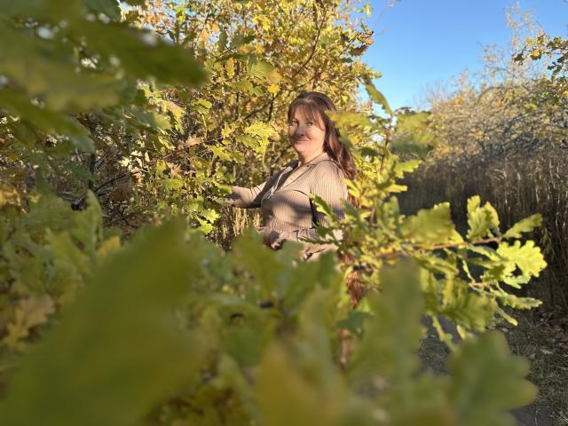 OVanessaHotly with brown hair, medium tits, in a beige top, standing amid green and yellow leaves, sunlight on her face, blue sky in background, nature setting.