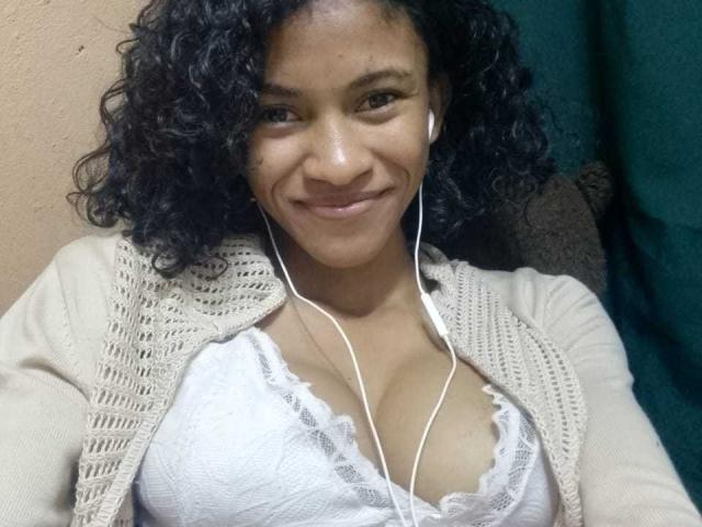 Alya69x with curly black hair, smiling, wearing white earbuds, a beige knit cardigan, and a lace-trimmed white bra, showing her medium-sized tits.