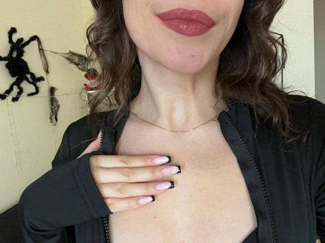 Sevillia's face, showing red lipstick and wavy brown hair. She's wearing a black zip-up, hand on chest, pink and black nails, and a thin gold necklace. Boobs visible.