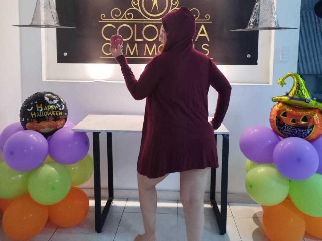BiancaWild in a red dress, hood up, holding a cup, standing in front of a table with colorful balloons and Halloween decorations, including a green witch hat.