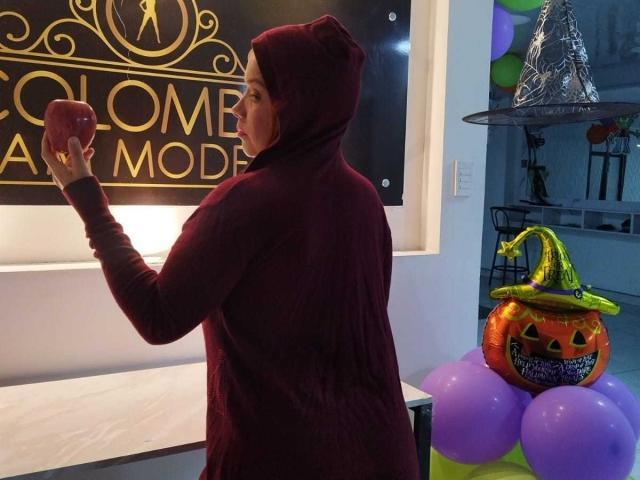 BiancaWild in a red hoodie, holding a pink cup, standing next to a Halloween pumpkin bucket with a green witch hat, and purple balloons. Her ass is slightly visible.