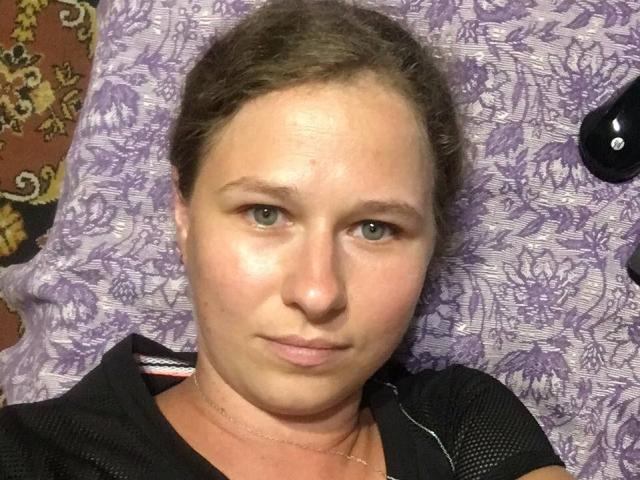 CurlyKsu has a slightly flushed face, light green eyes, and a subtle smirk. She's lying on a purple patterned pillow with a black button on the right.