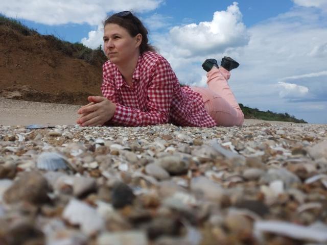 CurlyKsu is lying on a pebbled beach, wearing a red checkered shirt and pink pants, with her ass raised and legs bent, under a blue sky with clouds.
