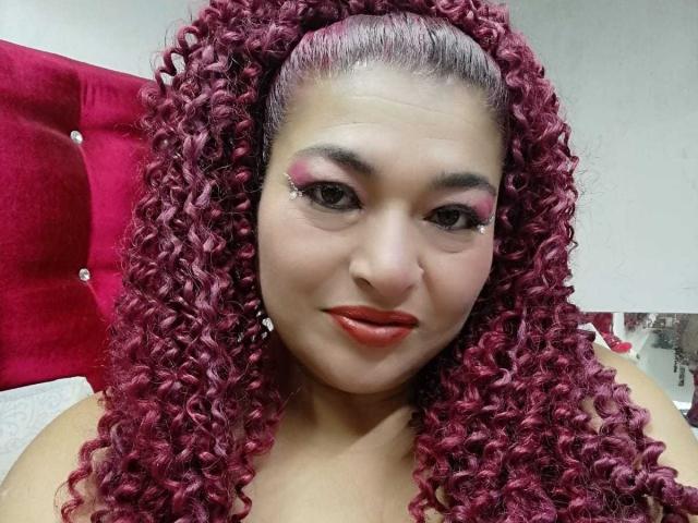 BriannaAngels with curly magenta hair, pink eyeshadow, and red lipstick, smiling at the camera, showing her tits slightly above the frame, in a bedroom with a red headboard.
