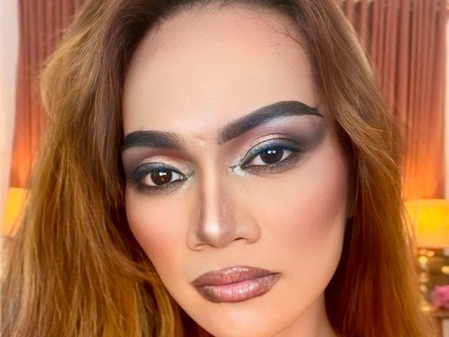 HugeCockJackie has a serious face with smoky eye makeup, thick eyebrows, and shiny pink lipstick. She has light brown, wavy hair and a fair skin tone.